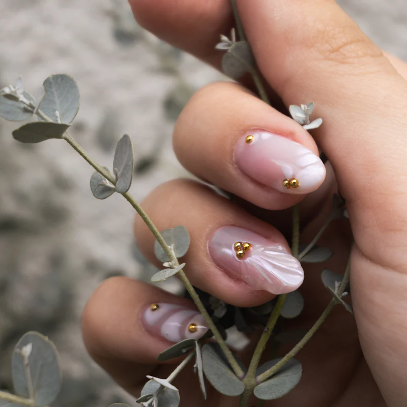 Milky pink nails with pearl and gold accents holding eucalyptus