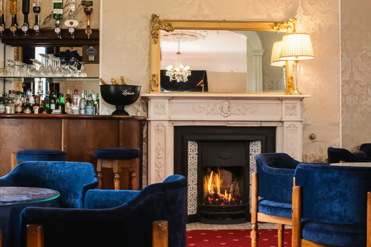 A warm lounge with an open fire, blue velvet armchairs and a small bar with a gilt mirror