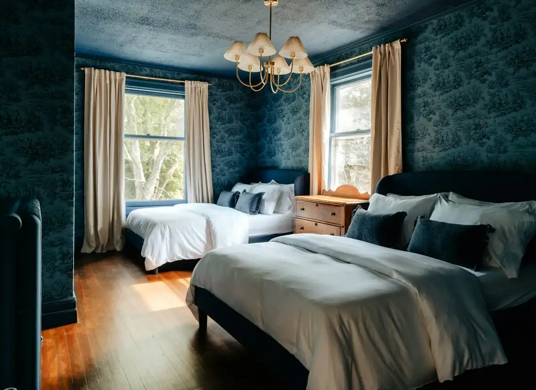 A bedroom with deep blue patterned wallpaper, a small chandelier and tall sash windows