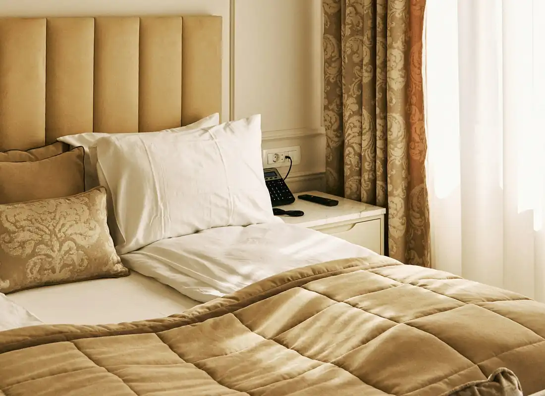 A light-filled bedroom with a padded headboard, a brass wall light and long patterned curtains