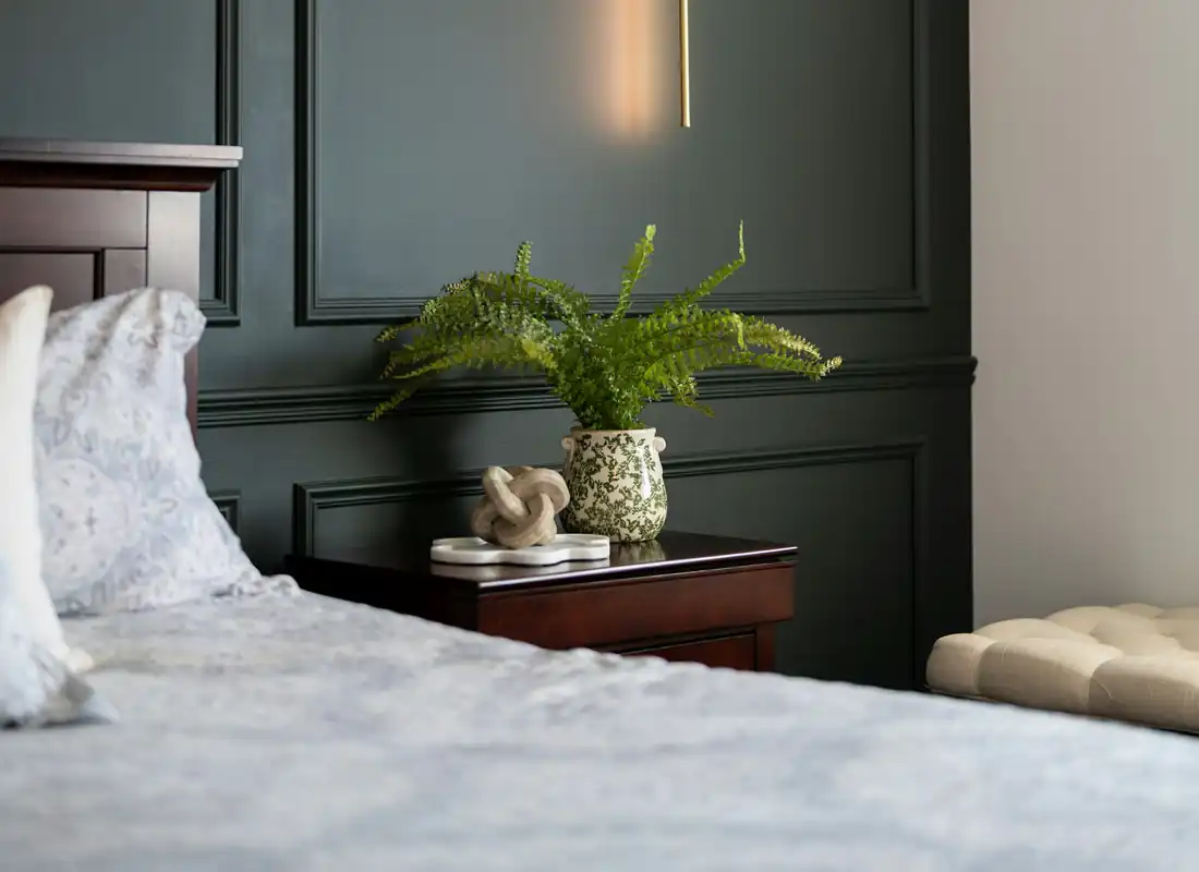 A calm bedroom with dark green panelled walls, a slim brass wall light and a fern on the bedside table