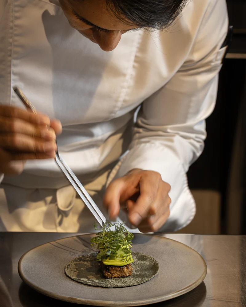 Chef in whites carefully garnishing a plated dish with herbs using tweezers