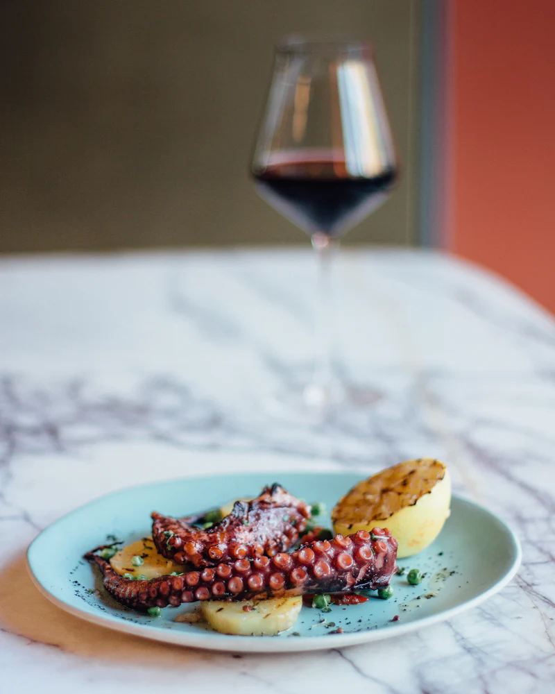 Charred octopus with potato on a pale blue plate next to a glass of red wine